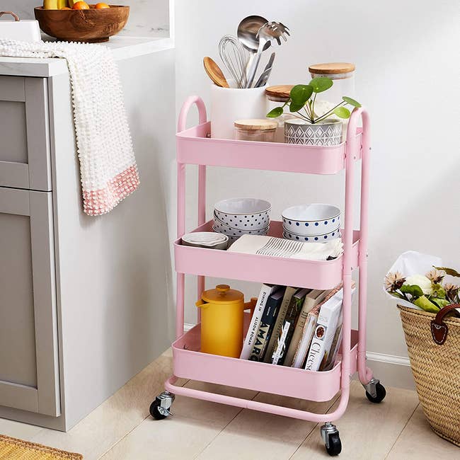 light pink compact utility cart with three shelves filled with cookbooks, kitchen utensils, and bowls