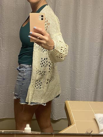 Person taking a mirror selfie wearing a crochet cardigan, green top, and denim shorts in a bathroom setting