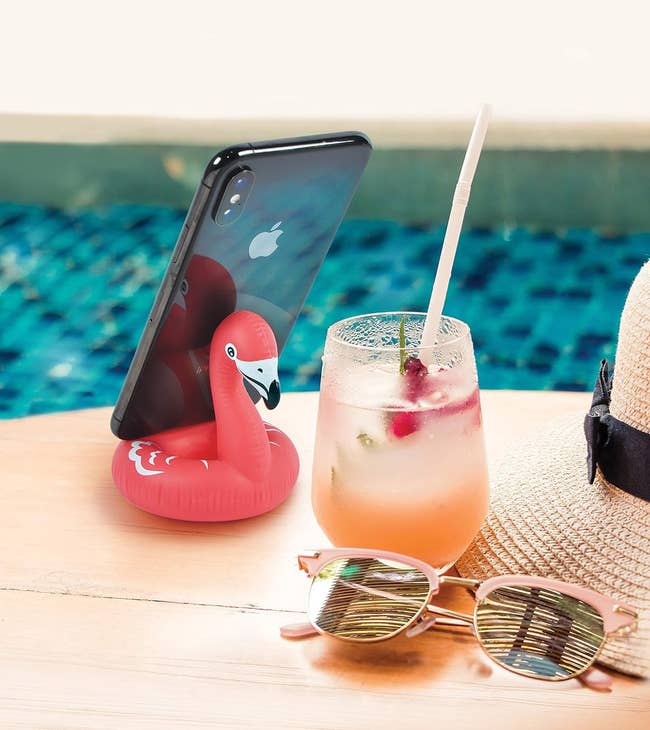 Smartphone on a mini flamingo holder next to a cocktail, sunglasses, and a sunhat by a pool