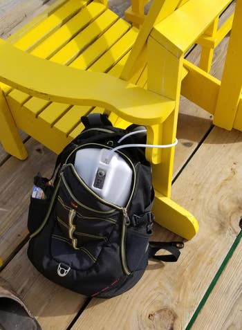 Black backpack holding a portable oxygen concentrator beside a yellowish woody seat connected a deck, with sunlit wood planks beneath
