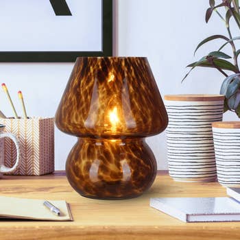 Stylish glass lamp with a textured surface, illuminated on a wooden desk with notepads and decorative cups nearby. Ideal for home decor shopping