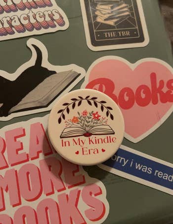 reviewer's Kindle covered in bookish stickers with a PopSocket that says In My Kindle Era