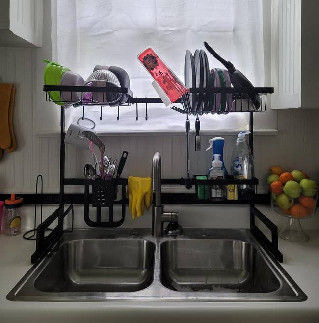 Kitchen sink with dish rack above holding various dishes and utensils, flanked by cleaning supplies and fruit bowl