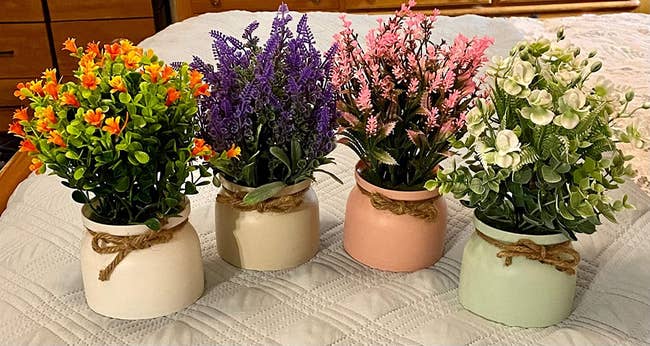 Reviewer image of orange, purple, pink, and white fake flowers inside small pots with rope tied around the neck sitting on top of blanket