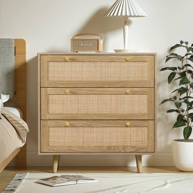 Wooden dresser with a woven front design, topped with a lamp and a small speaker, next to a potted plant. An open book lies nearby