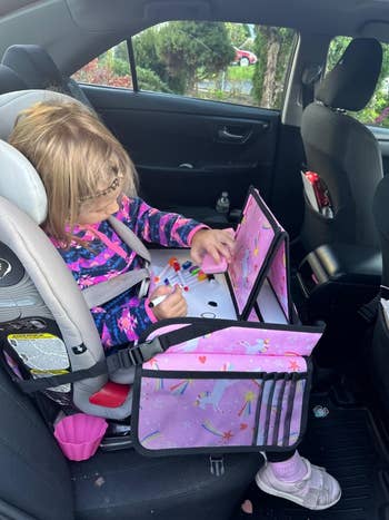 A child sits in a car seat while drawing on a car seat organizer tray