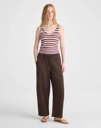 A person wearing a striped tank top, dark loose pants, and sandals, standing in a neutral pose, suitable for a fashion or shopping article