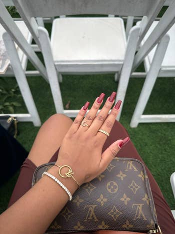 Hand with manicured nails, wearing gold rings and bracelets, placed on a designer purse. Elegant seating setup visible in the background