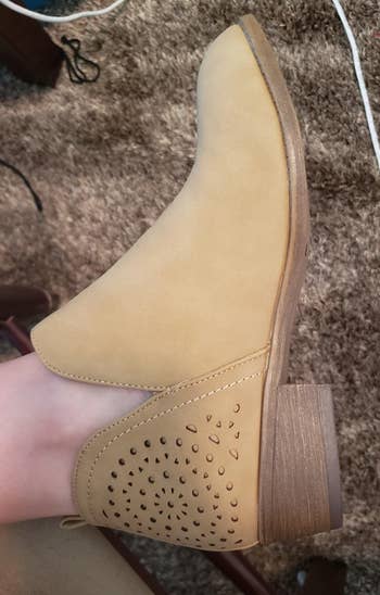 a reviewer wearing a light tan ankle boot with a short block heel and cutouts along the back heel 