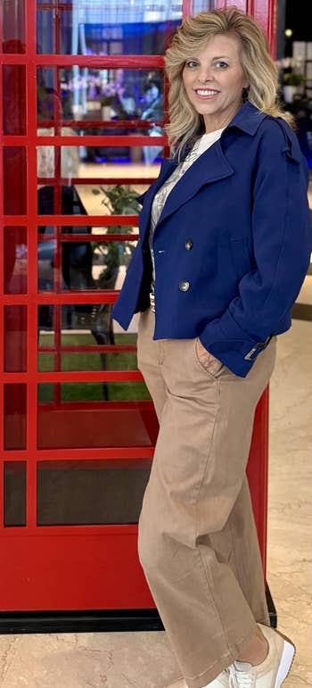 Person successful a bluish overgarment and beige pants stands adjacent to a classical reddish telephone booth indoors