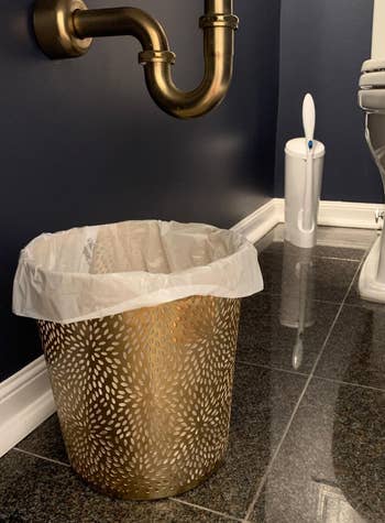 reviewer photo of the chrysanthemum patterned gold wastebasket in a bathroom