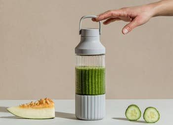 the bottle in grey with a thick green liquid inside sitting next to cucumbers and a piece of cantaloupe 