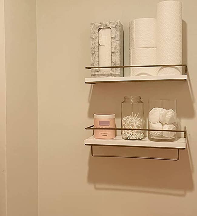 Reviewer image of two white shelves with golden bars hung on top of a toilet with toiletries lining each one