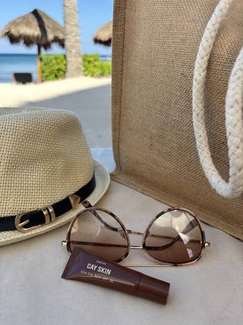  Straw chapeau  with a achromatic  band, tortoiseshell sunglasses, and Cay Skin SPF articulator  balm beside a woven tote connected  a sandy beach