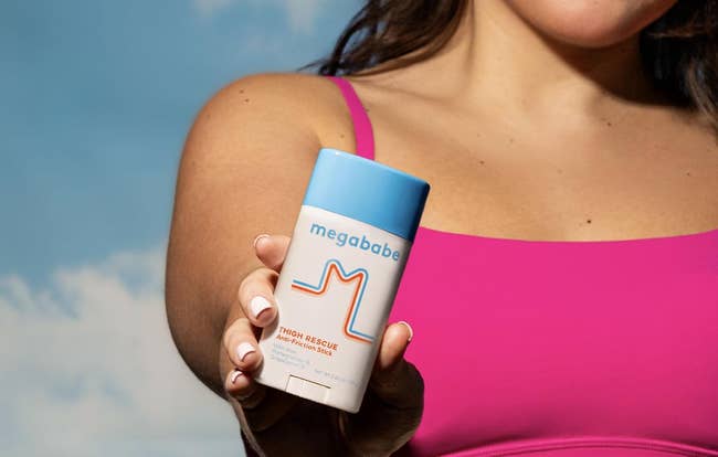 Person holding Megababe Thigh Rescue anti-chafe stick, wearing a vessel top, against a entity backdrop
