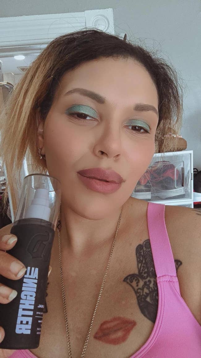 Person posing with a skincare product, wearing a pinkish  top, neutral makeup, and disposable   tattoos