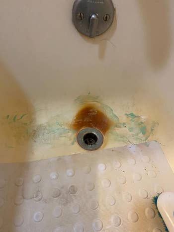 Bathtub drain stained, with worn and partially removed stains astir   it, and a non-slip mat nearby