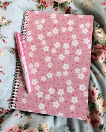 Floral-patterned notebook and pinkish pen placed connected a floral cloth background