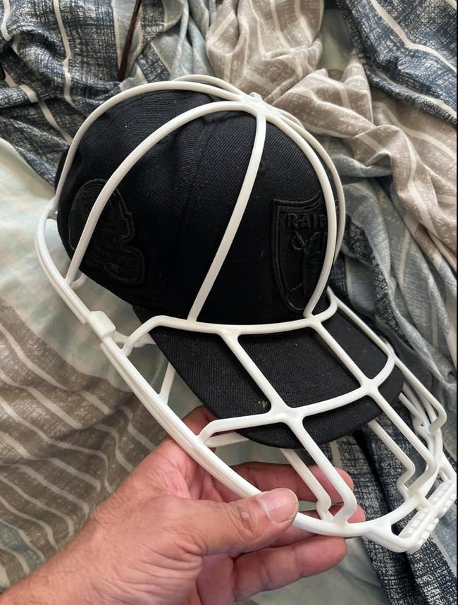 A hand holding a black cap fitted with a white 3D-printed frame shaped like a football helmet