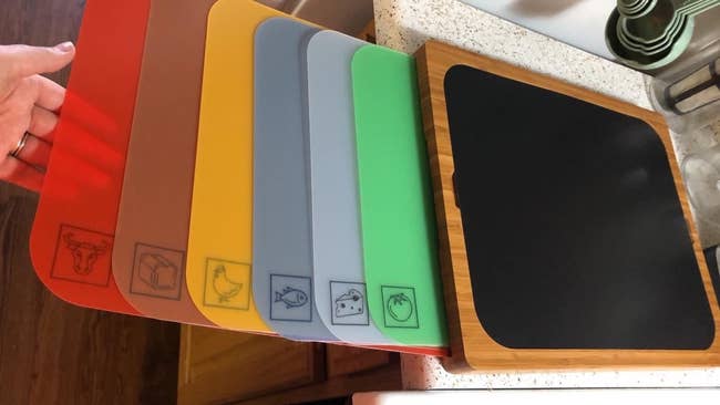 a reviewer photo of the bamboo cutting board on the counter with the reviewer holding the different-colored, labeled mats 