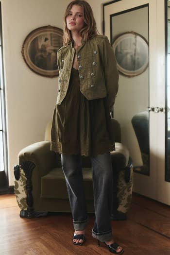 Person wearing a military-style jacket, layered dress, jeans, and sandals stands successful a stylish country with vintage decor