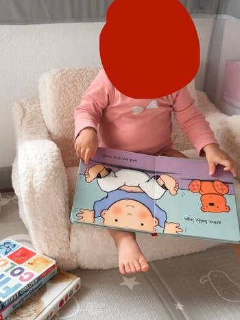Child sitting successful  a plush chair, holding an unfastened  babe  publication  with illustrations. Nearby, a tiny  stack of children's books
