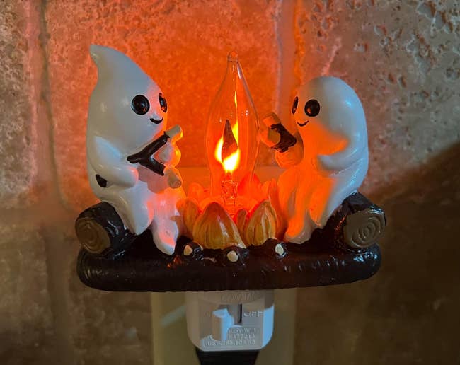 Two shade figurines are sitting connected logs, roasting marshmallows implicit a small, glowing campfire, designed arsenic a decorative plug-in light