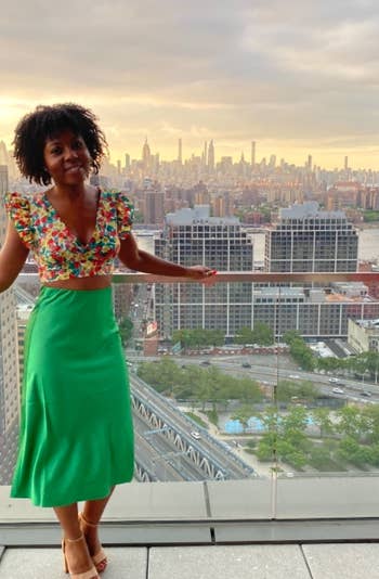 reviewer on balcony wearing a floral top and green skirt, city skyline in background