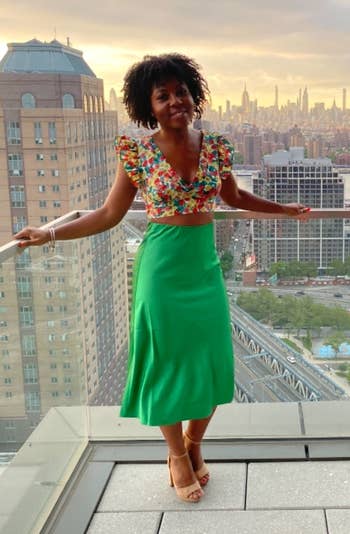 reviewer on balcony wearing a floral top and green skirt, city skyline in background