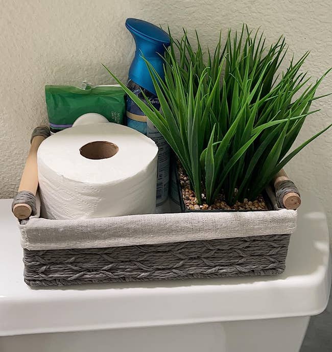 A woven handbasket with toilet paper, aerial freshener, and works connected a toilet tank
