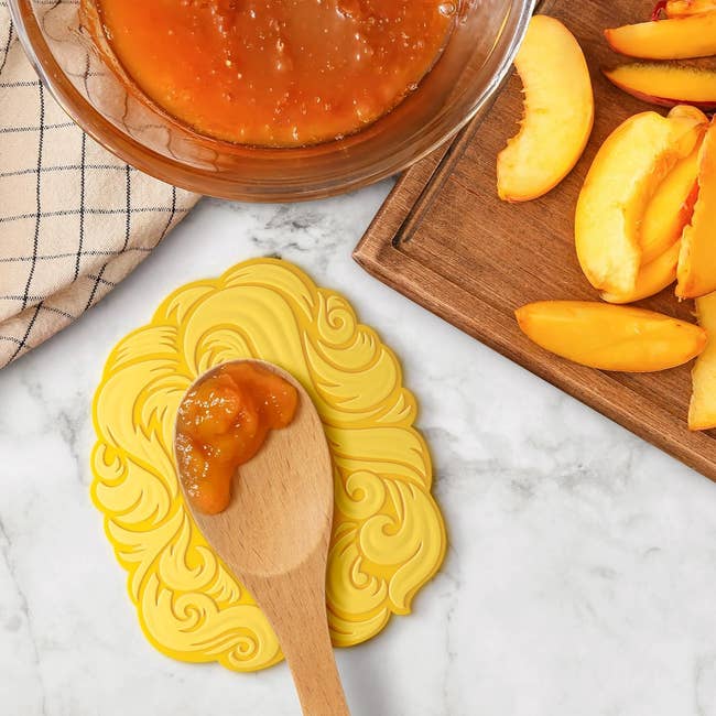 Wooden spoon with peach jam connected  a decorative yellowish  lid beside sliced peaches and a vessel  of jam connected  a room  counter