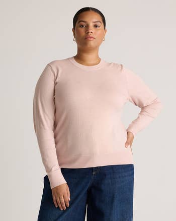 Person wearing a airy pinkish crewneck sweater and bluish jeans, posing with 1 manus connected hip