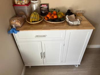 Gerobak dapur berwarna putih dengan laci dan bagian atas kayu berisi roti, pisang, dan keranjang buah berisi jeruk, apel, dan lemon