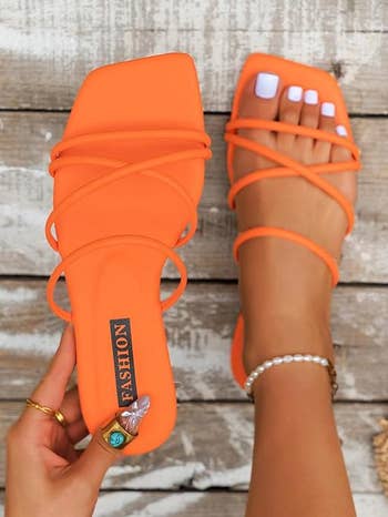 model wearing one orange strappy sandal and holding the other
