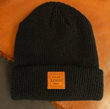 Beanie in knit black with small brown leather square in center that reads 