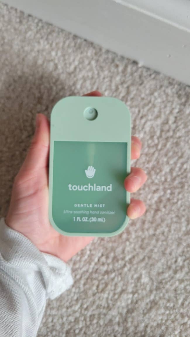 Person holding a vessel of Touchland gentle mist manus sanitizer, 1 fl oz (30 mL), against a neutral background