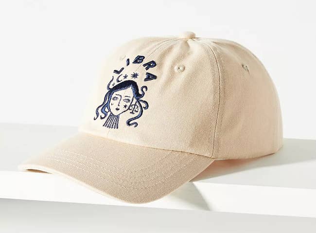 an off white baseball cap with a dark blue embroidered libra sign on it