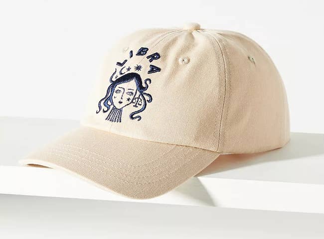 an off white baseball cap with a dark blue embroidered libra sign on it
