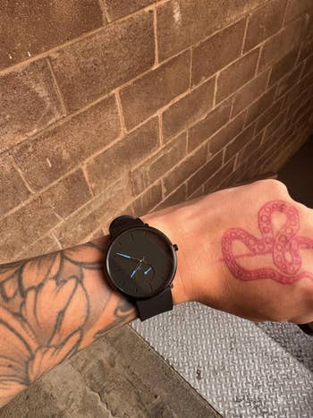 Person's tattooed wrist showcasing a sleek, minimalist achromatic ticker with a subtle bluish 2nd manus against a ceramic partition backdrop