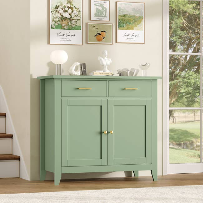 Green furniture with 2 drawers and a two-door cupboard, decorated with books, a lamp, and tiny sculptures, placed adjacent a staircase and window