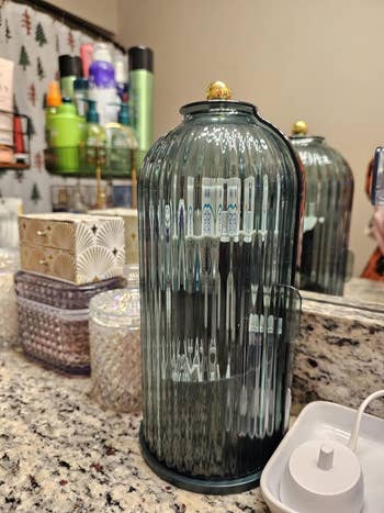 Glass instrumentality  with vertical ridges connected  a bath  counter, surrounded by toiletries and decorative boxes