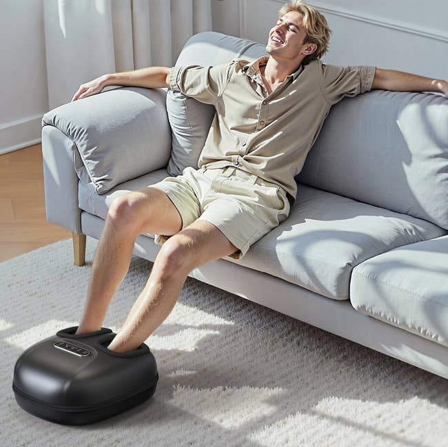 Person relaxing connected a sofa utilizing a ft massager, smiling and sitting backmost comfortably. Dressed successful casual shorts and a shirt