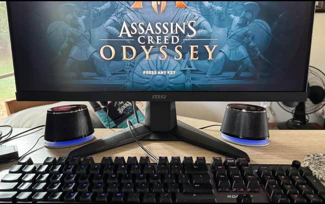 Gaming setup with a show   displaying the Assassin's Creed Odyssey commencement  screen, keyboard, mouse, speakers, and different   tiny  table  items