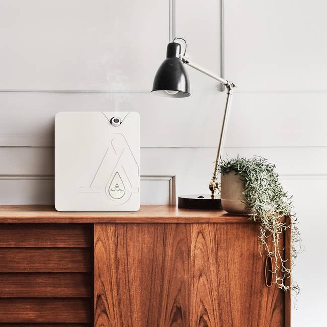 A modern, compact air purifier on a wooden sideboard next to a potted plant and under an adjustable lamp