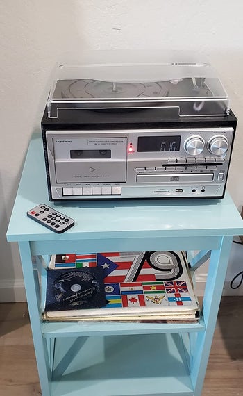 Silver turntable with cassette player, CD player, and radio with customizable dials, volume control, and buttons, and a clear cover on top of light blue side table