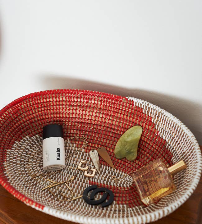 The oval red and white woven bowl with jewelry, a gua sha, perfume, and a cosmetics bottle in it