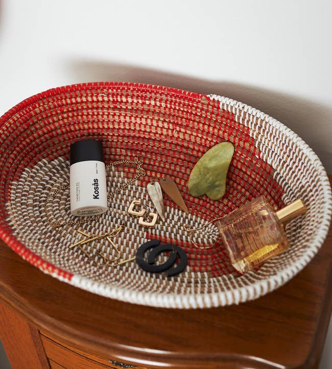 The oval red and white woven bowl with jewelry, a gua sha, perfume, and a cosmetics bottle in it