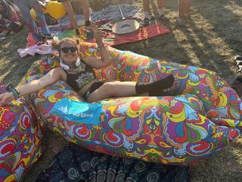 Person lounging on a colorful inflatable lounger at an outdoor event, wearing casual festival attire with sunglasses and headphones