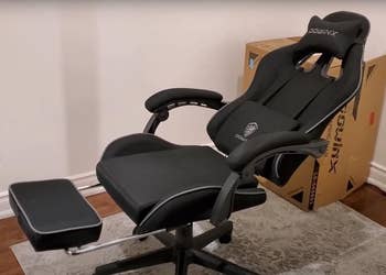Ergonomic gaming chair with armrests, reclined, and featuring a foldable leg rest, set against a small rug and a cardboard box in the background