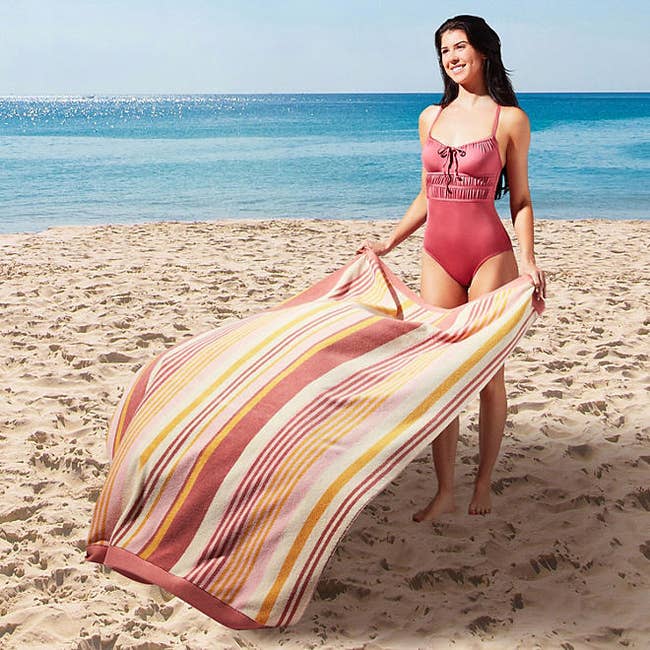 Woman in a one-piece swimsuit holding a striped beach towel on the sand